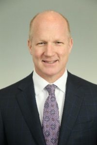 Clymer Bardsley, fair skin, bald, smiling, wearing a white dress shirt with purple tie and black suit coat